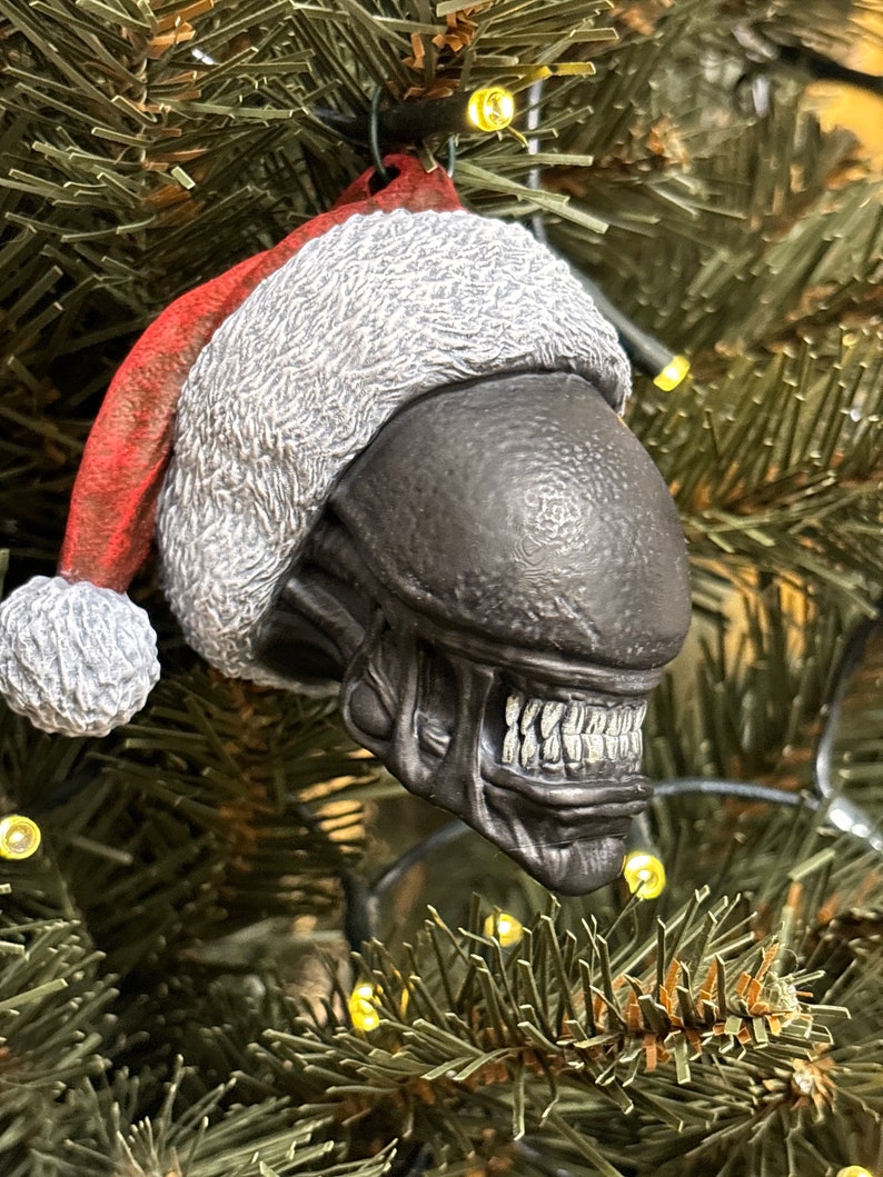 May include: A black alien Christmas ornament wearing a red and white Santa hat. The ornament is detailed with a textured surface and visible teeth.