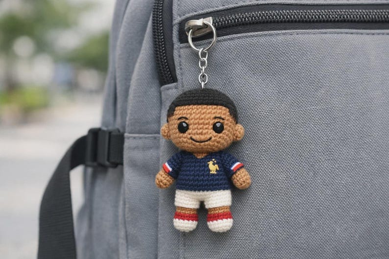 May include: Crocheted keychain featuring a small doll in a navy blue jersey with red and white accents, white shorts, and red-striped socks. The doll has black hair and is attached to a silver keychain, hanging against a gray backpack.