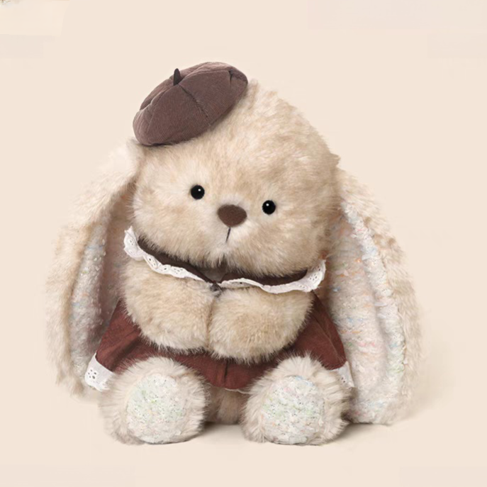 Brown hat rabbit plush toy wearing lace-trimmed dress, beige stuffed animal sitting on gift box