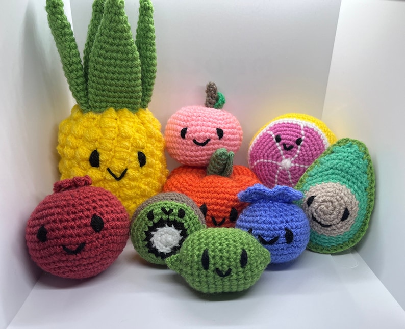 May include: A collection of crocheted fruit and vegetable toys. The toys are all different colors and shapes, including a pineapple, a red pomegranate, a kiwi, a green avocado, a blue blueberry, a pink grapefruit, and an orange pumpkin. The toys have happy faces with black eyes and mouths.