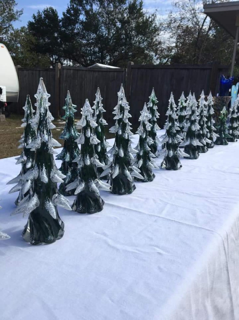 May include: A row of green glass Christmas trees with white glitter on a white tablecloth. The trees are arranged in a line from left to right, with the tallest tree on the left and the shortest tree on the right.
