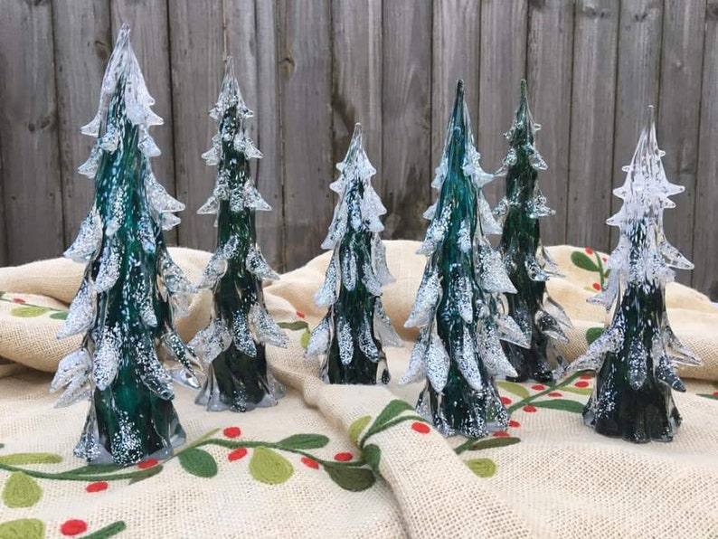 May include: Seven glass Christmas trees in various sizes, all with a frosted white finish and a green base. The trees are arranged on a beige burlap surface with a green and red floral pattern.