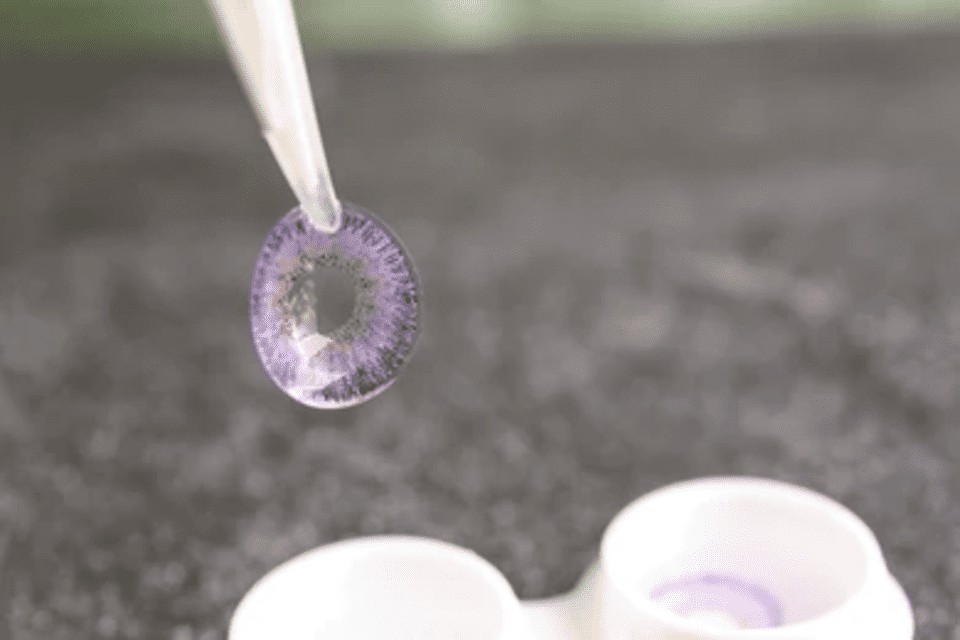 A pair of tweezers holding a purple colored contact lens above its case.