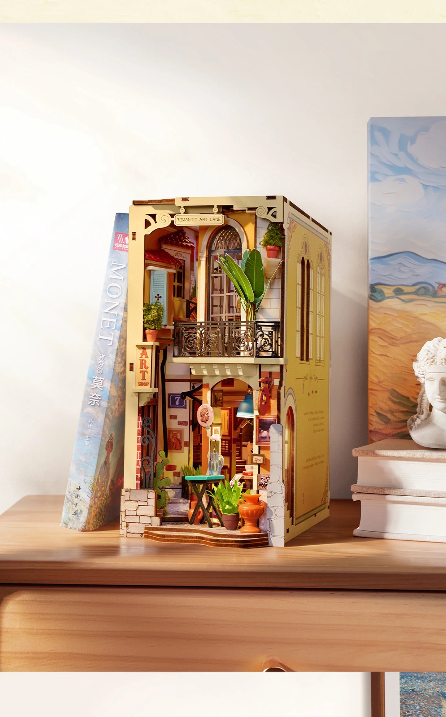 Rolife miniature art lane diorama with arched window, balcony, plants and warm interior lighting, displayed on a wooden surface between a Monet book and a small bust.