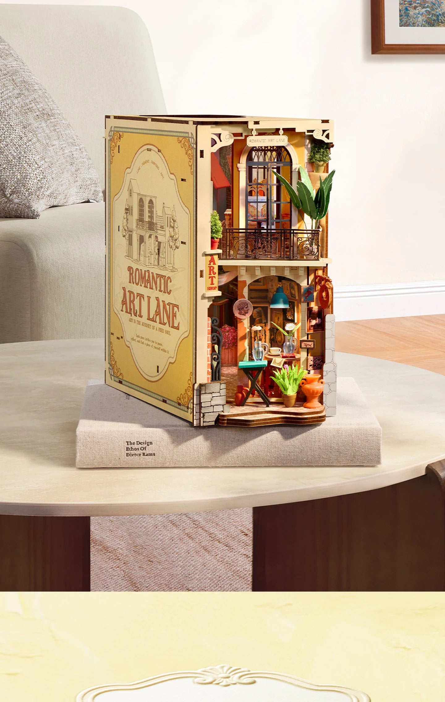 Rolife Romantic Art Lane miniature two-storey art shop diorama on a round table, showing a balcony, arched window, plants, caf&eacute; table and pottery, with a printed side panel.