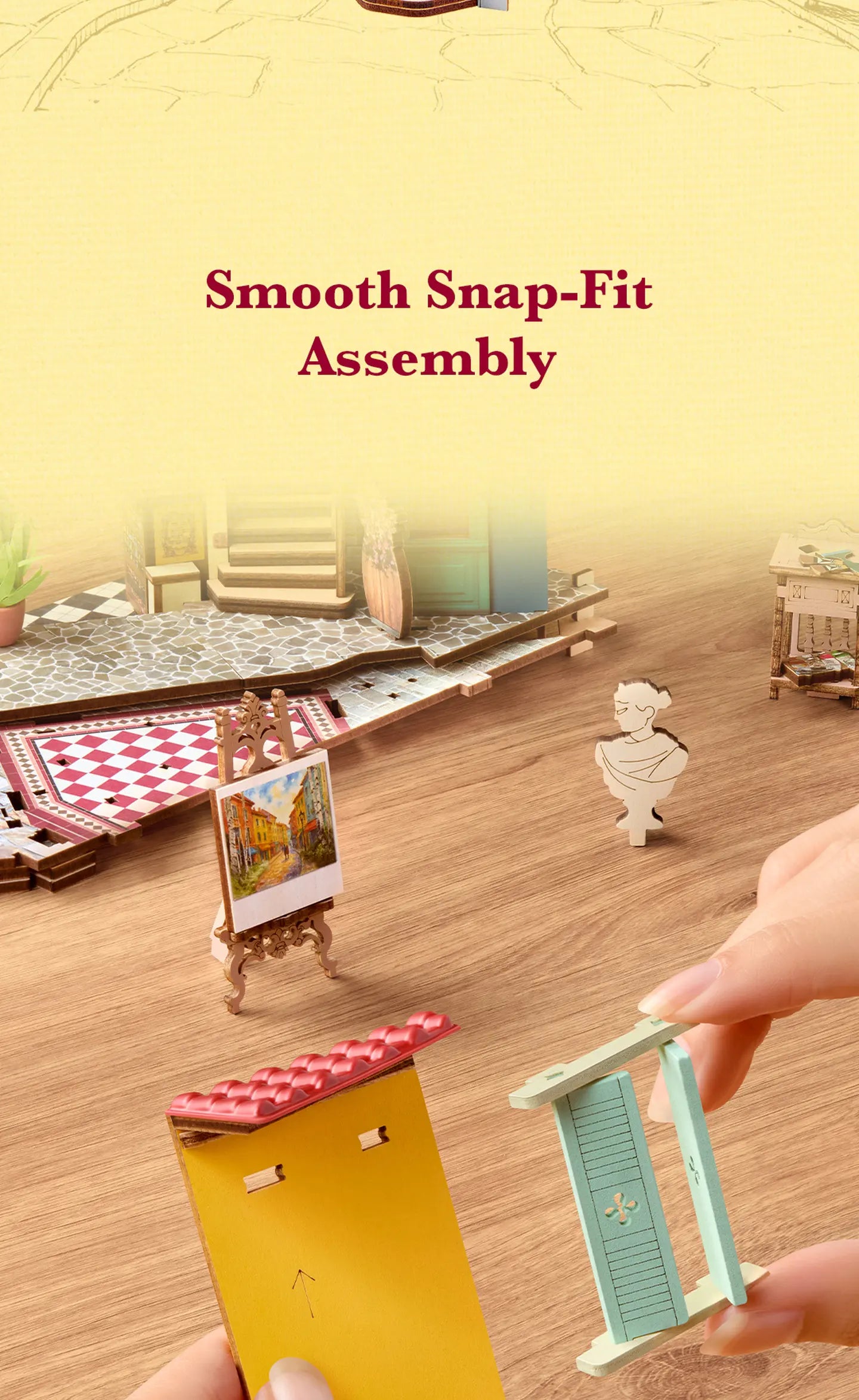 Hands assembling wooden miniature shop parts on a wooden table, with a painted easel, patterned floor and small cutout figure nearby.