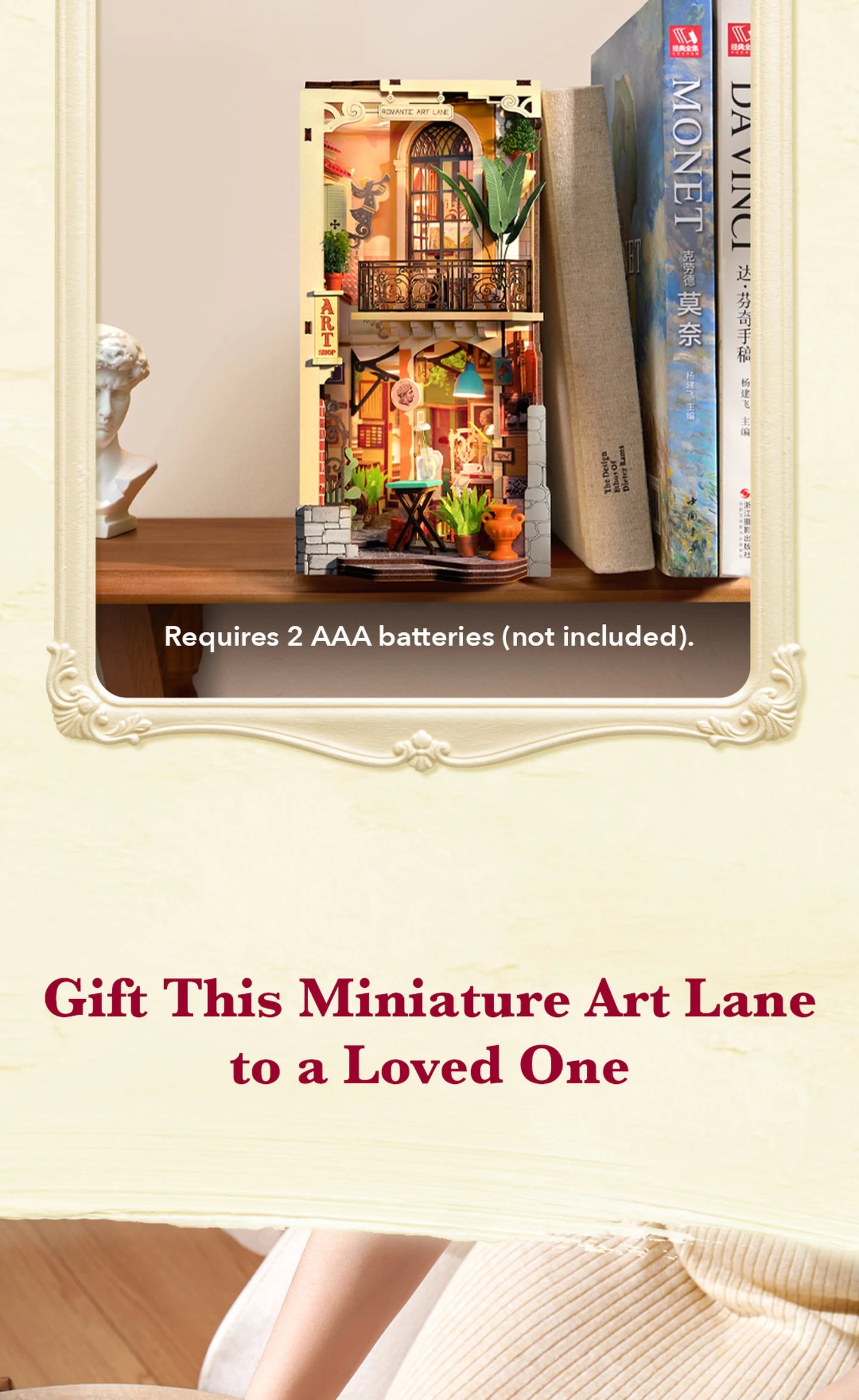 Miniature two-storey art lane diorama with balcony, plants and warm lighting, displayed between books on a wooden shelf beside a white bust; text notes 2 AAA batteries required.