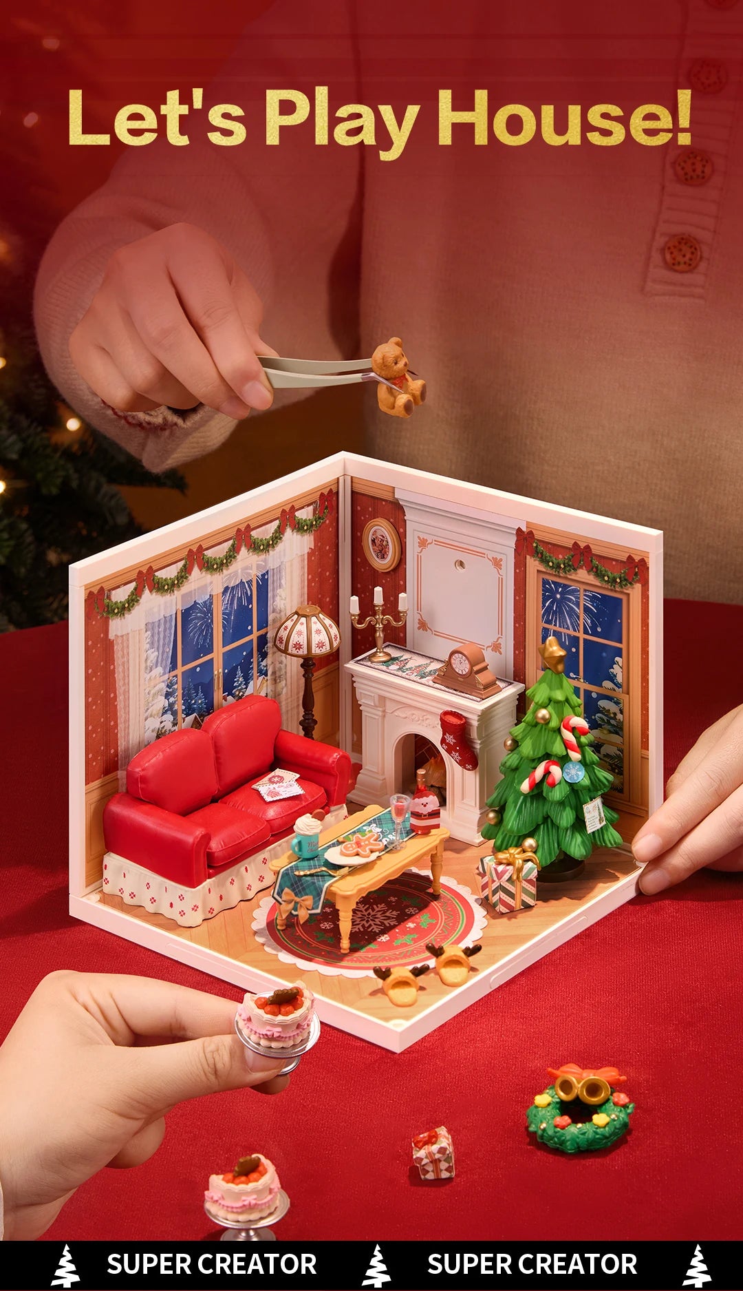 Hands using tweezers to place a miniature teddy bear into the assembled Christmas Eve room box, with loose cake, wreath and gift pieces on a red surface.
