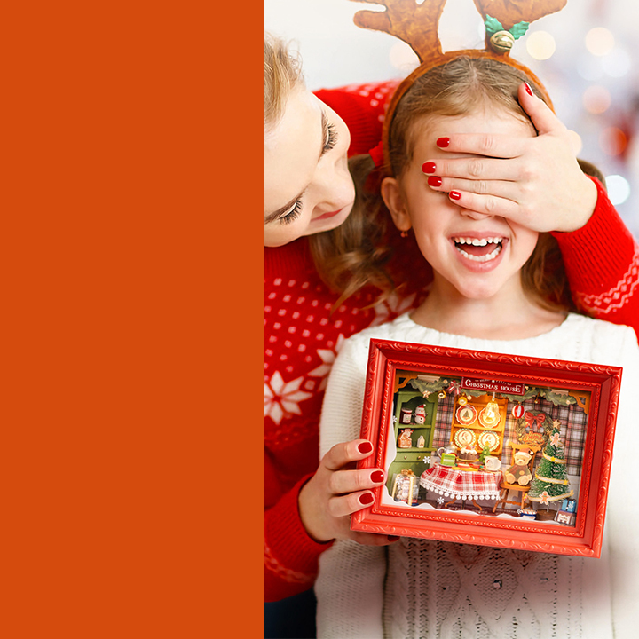 Mother covering a child's eyes while the child holds a Christmas miniature house