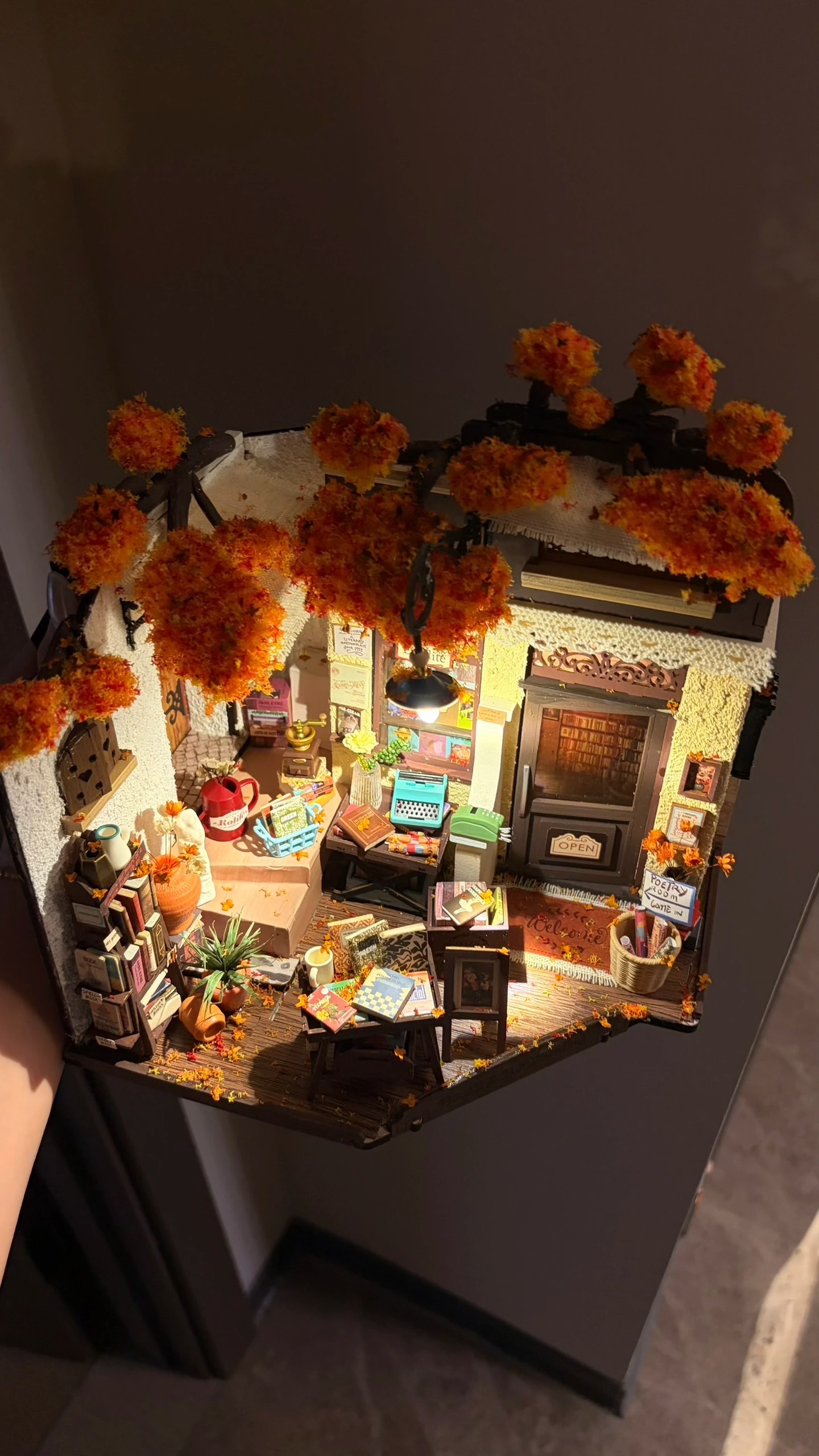 Rolife Community maker's Corner Librairie DG164 encadrée par des troncs d'arbres faits à la main, un feuillage orange, des feuilles d'automne éparpillées et un éclairage intérieur chaleureux.