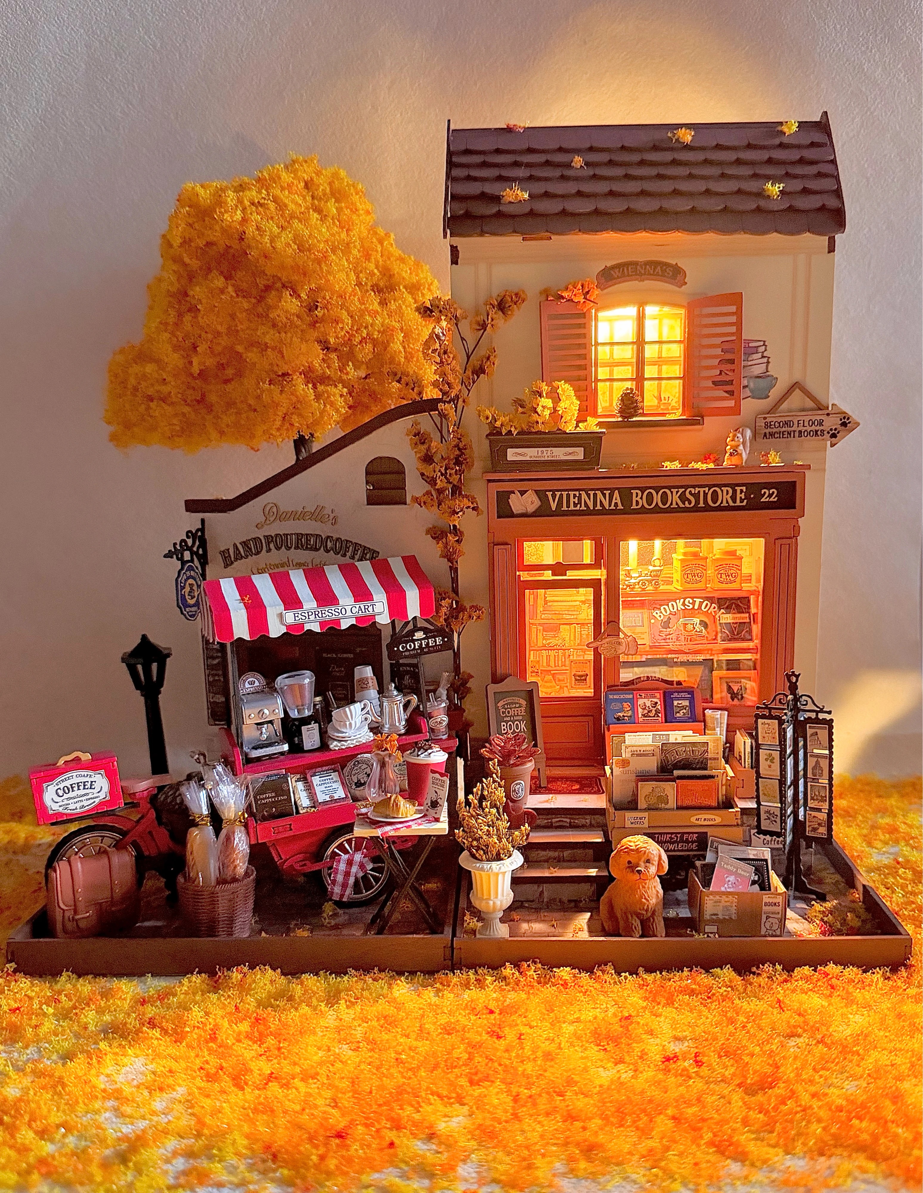 Librairie de Vienne DG183 de la communauté Rolife, stylée avec un chariot à café, un arbre d'automne doré, un tapis de feuilles orange, des figurines et une lumière chaleureuse.