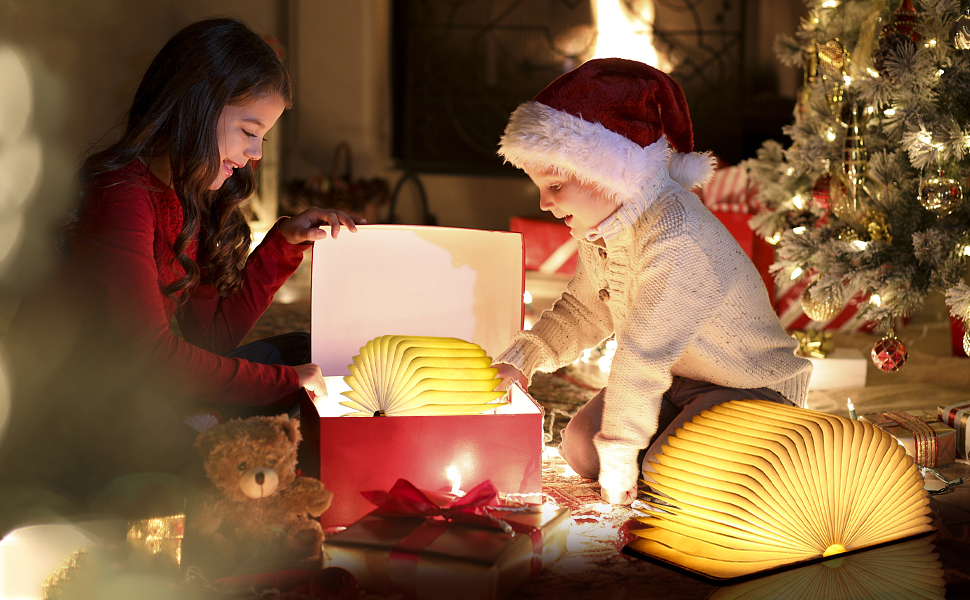 Two people opening a glowing book-shaped lamp near a Christmas tree. The lamp emits warm yellow light, creating a cozy atmosphere.