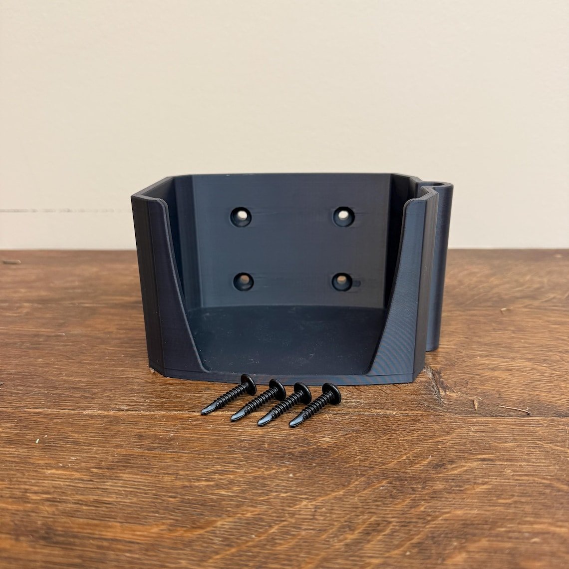 May include: A black plastic wall mount with four screw holes and a small lip on the side. Four black screws are in front of the mount.