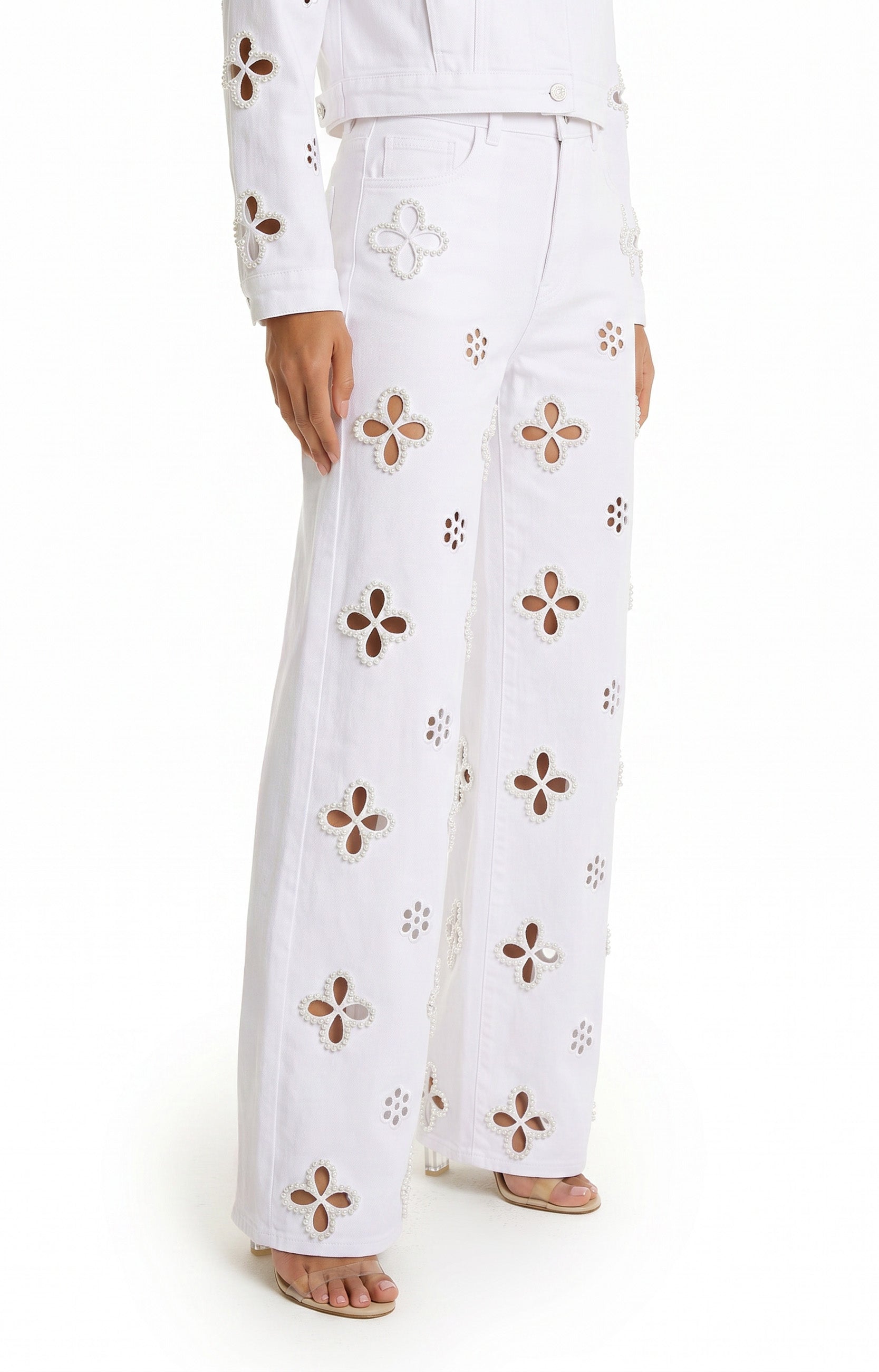 The lower half of a person is shown wearing the Kaia Pearl High Rise Jean in white stretch denim with wide legs and large floral cutout designs, paired with a matching white jacket, against a white background.