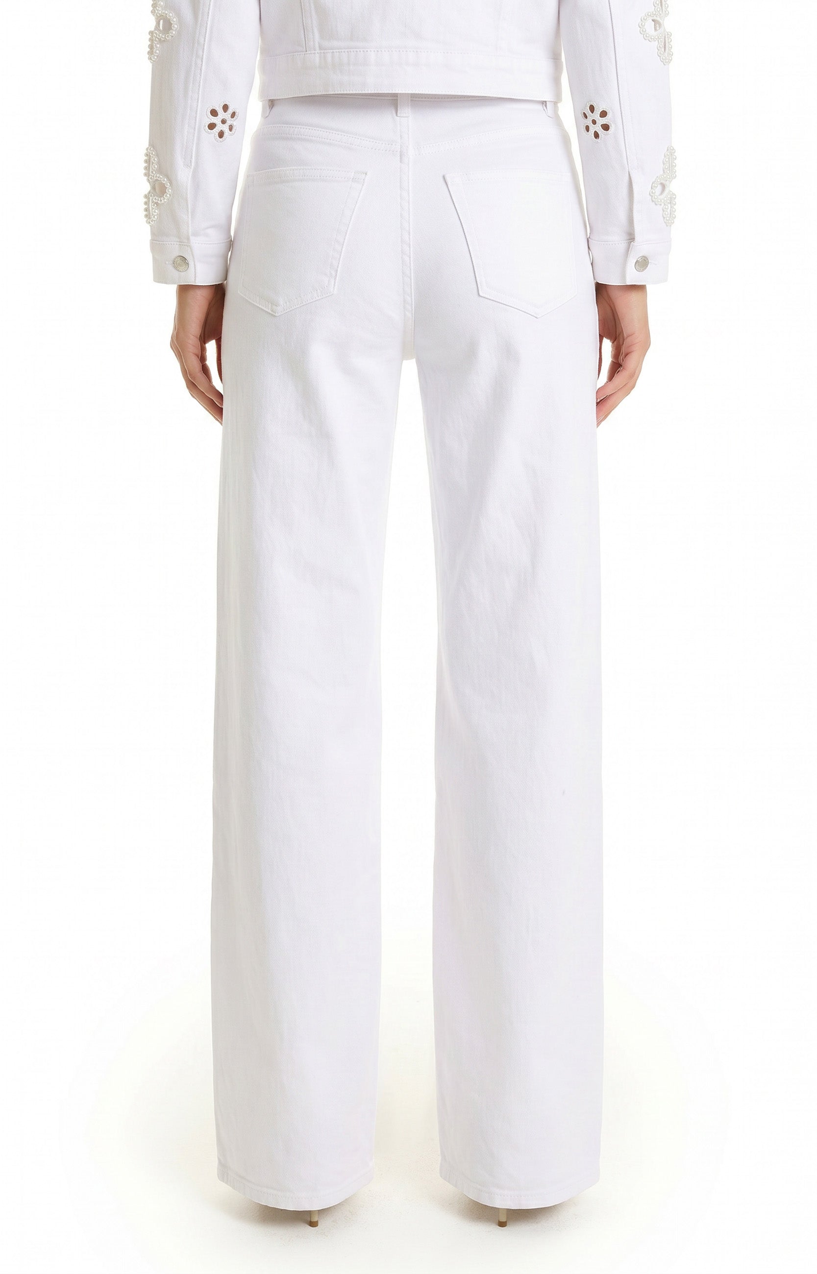 A person’s lower body and torso are visible, wearing the Kaia Pearl High Rise Jean in white stretch denim. They are facing away, styled with a white jacket featuring decorative floral sleeve patterns.