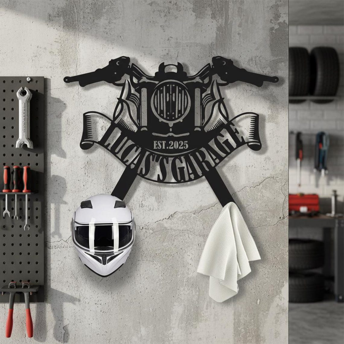 May include: Black metal motorcycle handlebar-themed wall decor with the text "LUCAS'S GARAGE EST. 2025". A white motorcycle helmet and a white cloth hang below the sign. The sign is mounted on a gray concrete wall.