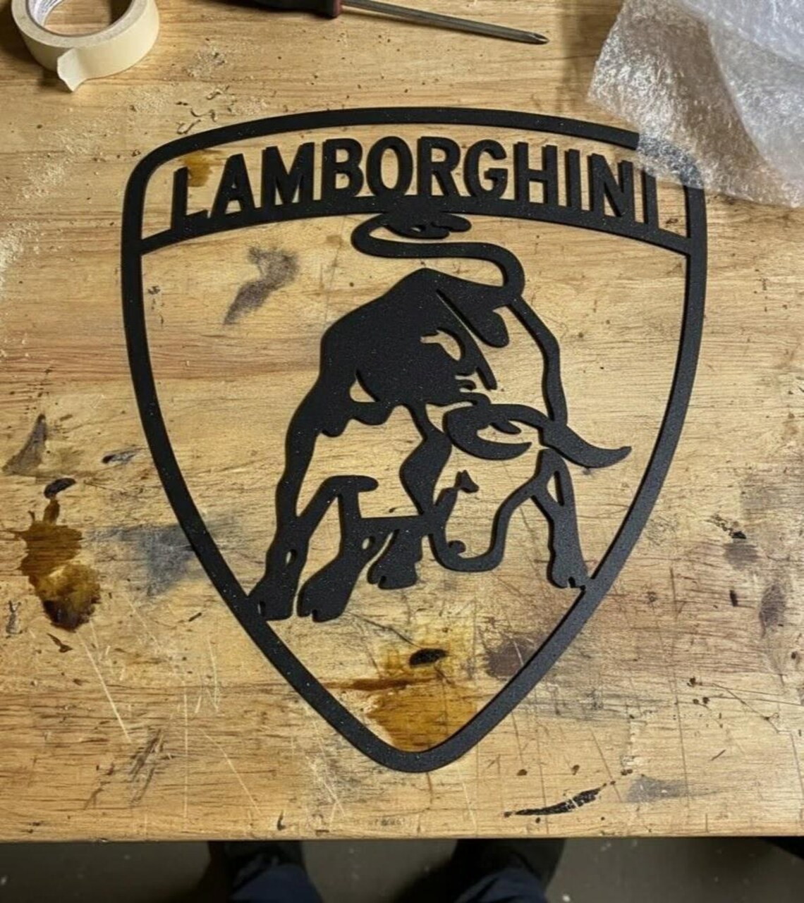 May include: Black metal Lamborghini logo in a shield shape. The logo features the word "LAMBORGHINI" at the top and a silhouette of a charging bull in the center. The logo is likely a decorative item or sign.