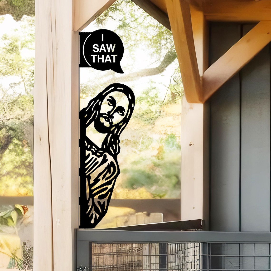 May include: Black metal cutout of Jesus peeking around a corner. Above, a speech bubble reads "I SAW THAT". The artwork is mounted on a wooden structure, with a blurred outdoor background visible through a window.