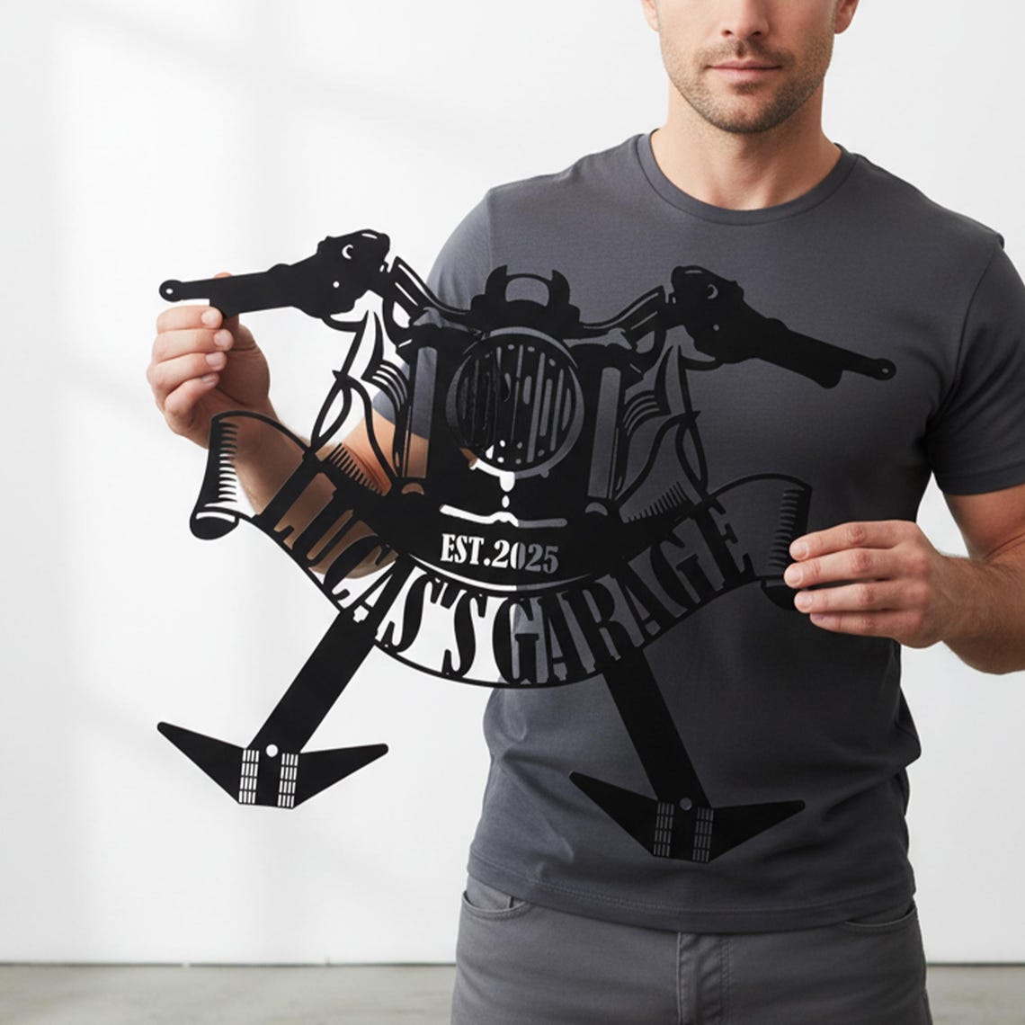 May include: Black metal motorcycle-themed wall art. The design features a motorcycle handlebar, headlight, and banner with the text "LUCAS GARAGE EST. 2025". The art piece is held up by a person.