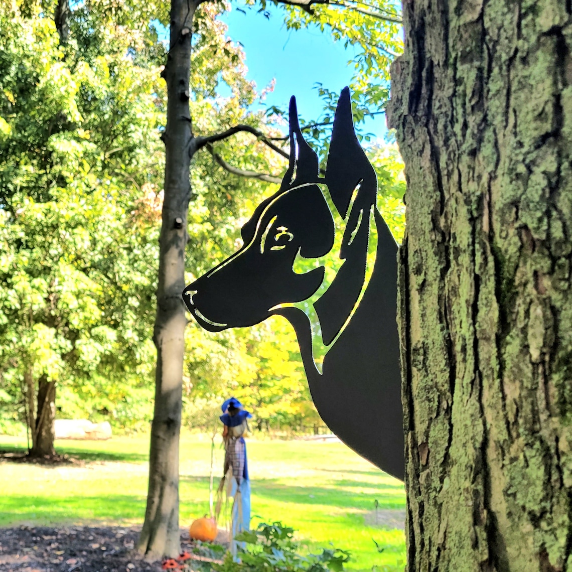 May include: A black metal silhouette of a dog's head is mounted on a tree trunk. The dog's head is facing to the left and the silhouette is cut out in a detailed design. The tree trunk is brown and has a rough texture.