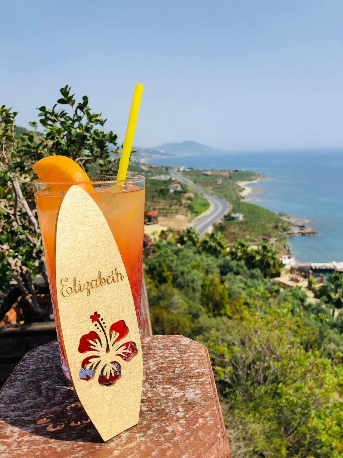 May include: A wooden surfboard drink stirrer with a red hibiscus flower cutout and the name 'Elizabeth' engraved on it. The stirrer is in a glass of orange and pink drink with a yellow straw. The drink is on a wooden table with a view of a coastline in the background.