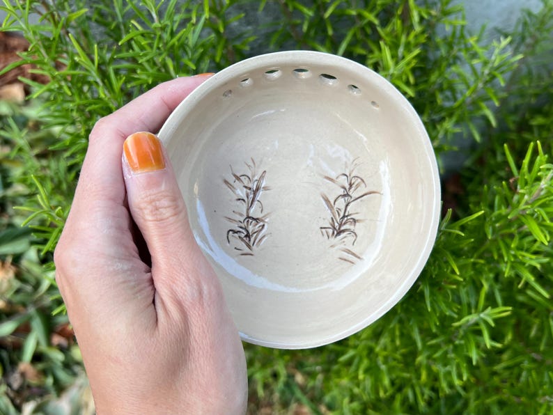 May include: A white ceramic bowl with a brown floral design. The bowl has small holes around the rim and a slightly raised lip.