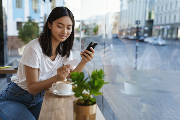 Free photo asian modern girl in a cafe, drink coffee, enters the credit card number in smartphone.