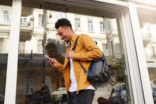 Free photo phortrait of young cheerful dark skinned guy in yellow shirt walking down the street , holds telephone, chatting with friends, broadly smiling, looks happy.