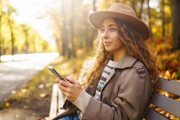 Photo young smiling woman sitting on bench in autumn park using phone in stylish clothes happy mood