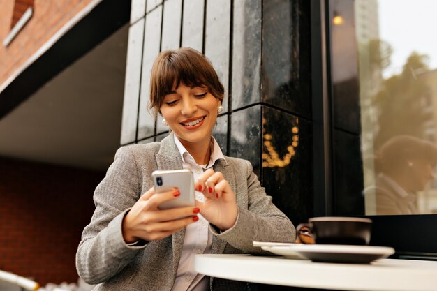 Free photo adorable young brunette lady in grey jacket and white shirt using smartphone and works in laptop