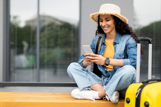 Photo cheerful young lady tourist using smartphone sitting on bench outdoors
