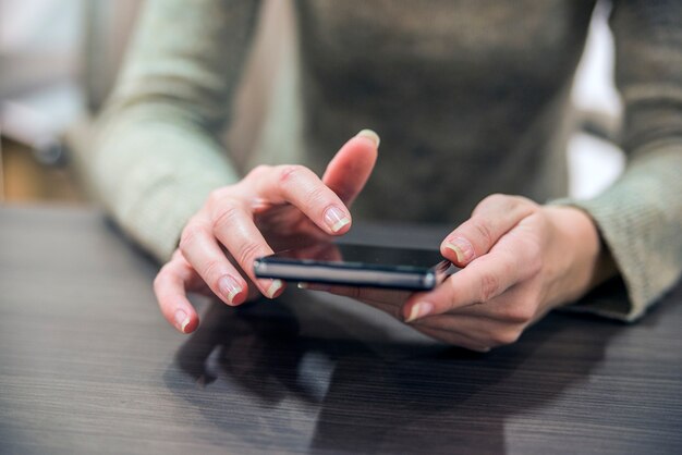 Free photo woman holding a smartphone on the wooden table. close-up hands of the girl, sitting at the wooden table, in one hand is smartphone. businesswoman surfing the internet on smartphone.