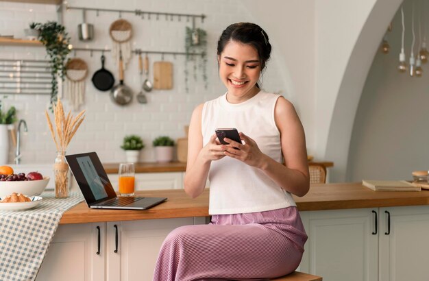 Free photo smiling young asian woman using mobile phone while sitting in kitchen room at home with laptop