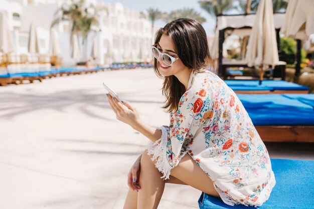 Photo gratuite portrait d'une fille brune avec des cheveux raides envoyant un message tout en se reposant dans une station balnéaire. Charmante dame en tenue à imprimé floral et lunettes de soleil assise sur un lit de jour sous le soleil tropical et souriant