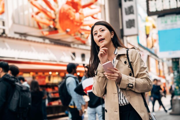 Photo d'une élégante fille asiatique utilisant son téléphone pour vérifier la bonne direction pour trouver un hôtel lors d'un voyage d'affaires au Japon. jeune femme tenant un téléphone portable utilisant une application de carte en ligne dotonbori osaka par une journée ensoleillée à l'extérieur