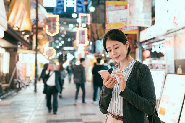 Photo d'une sortie nocturne d'une dame japonaise dans la rue à Tsutenkaku. Femme cherchant une application de carte en ligne sur son téléphone mobile en ligne la nuit. Jeune voyageur utilisant son téléphone portable pour trouver un restaurant pour le dîner.