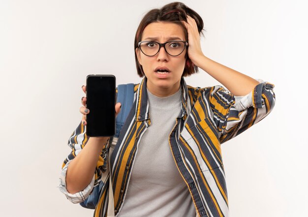 Free photo anxious young student girl wearing glasses and back bag showing mobile phone putting hand on head isolated on white