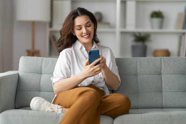 Photo happy casual beautiful woman is using a phone sitting on a sofa at home