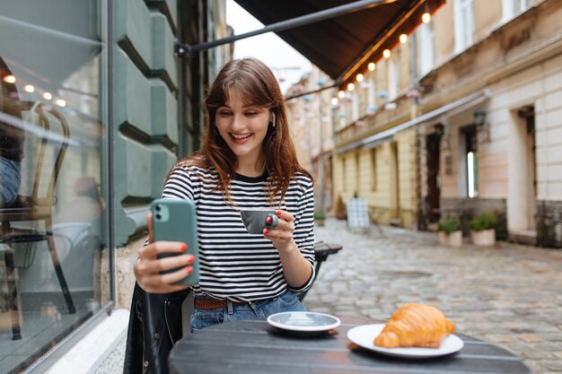 Foto de una mujer agradable vestida casualmente con auriculares inalámbricos bebiendo café y sosteniendo el teléfono celular