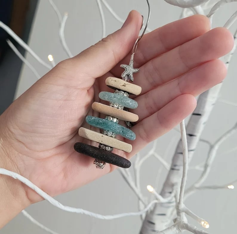 May include: A handmade Christmas tree ornament. The ornament is constructed from various colors of sea glass, including beige, light blue, and dark brown. The sea glass pieces are stacked and connected with silver-colored wire and small, clear rhinestones. A silver starfish tops the tree.