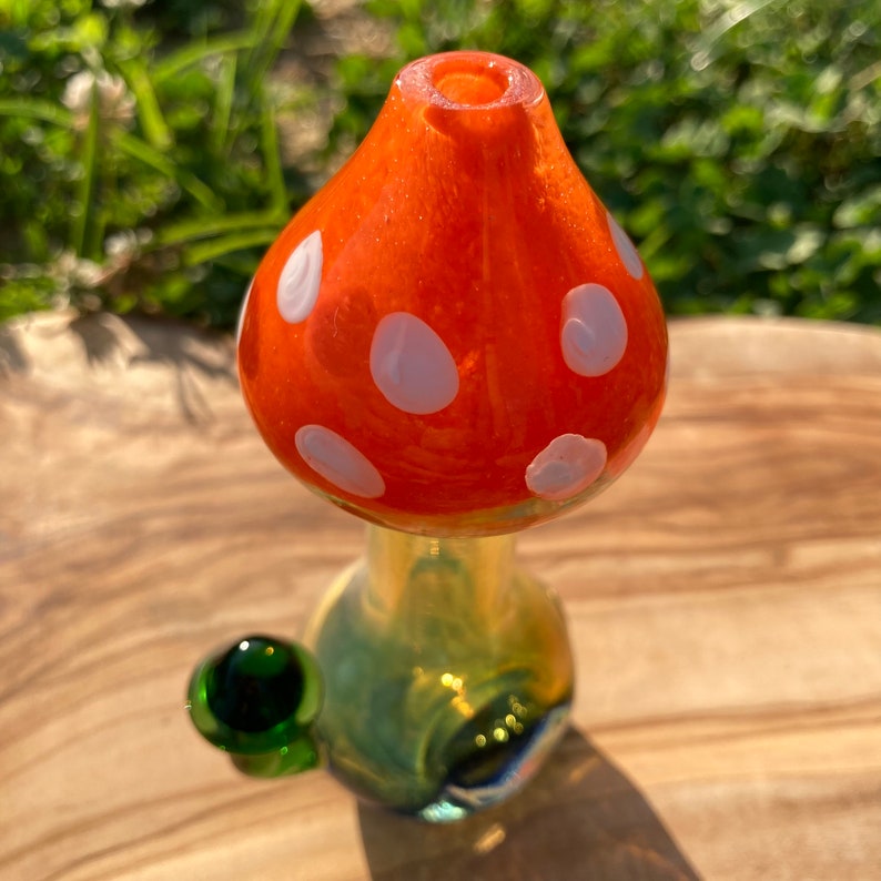 May include: A glass mushroom-shaped pipe with a green glass bowl. The top of the pipe is orange with white polka dots. The bottom of the pipe is green and yellow.