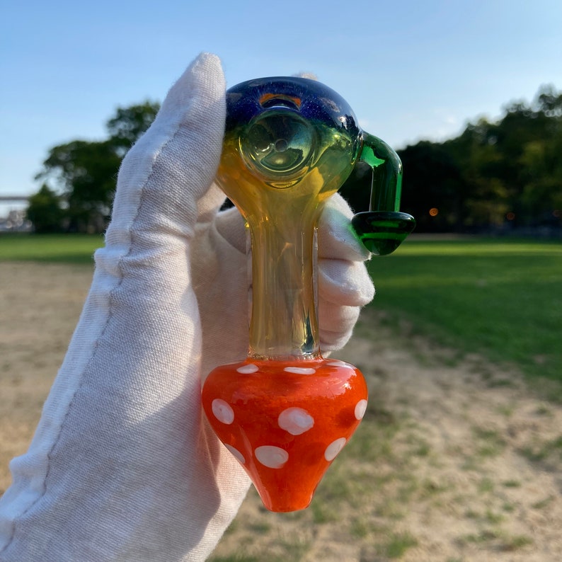 May include: A glass pipe with a mushroom-shaped bowl. The bowl is orange with white polka dots. The pipe is made of clear glass with blue, green, and yellow accents.