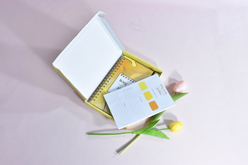 yellow journal and notepad inside of yellow box. notepad is slightly to the left. pink and yellow flowers are next to the box that is holding the journal and notepad.