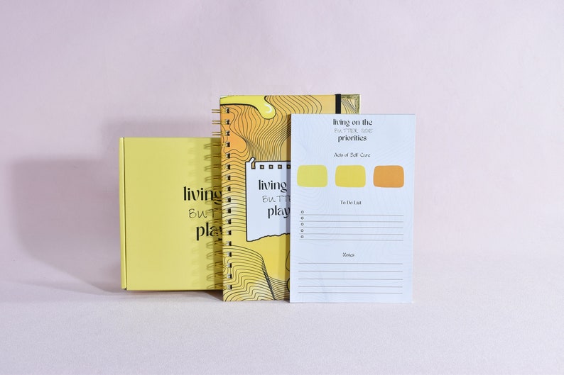 image of yellow journal with gold corners and gold binding. white notepad with yellow details and lines to write information. yellow box behind the journal and notepad. all items are standing up right.