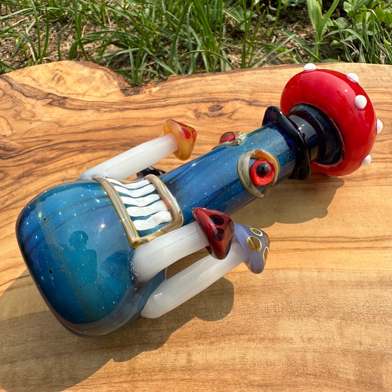 May include: A blue glass water pipe with a red and white mushroom top. The pipe has white and gold accents and features a mushroom design with red and white details. The pipe is sitting on a wooden surface.