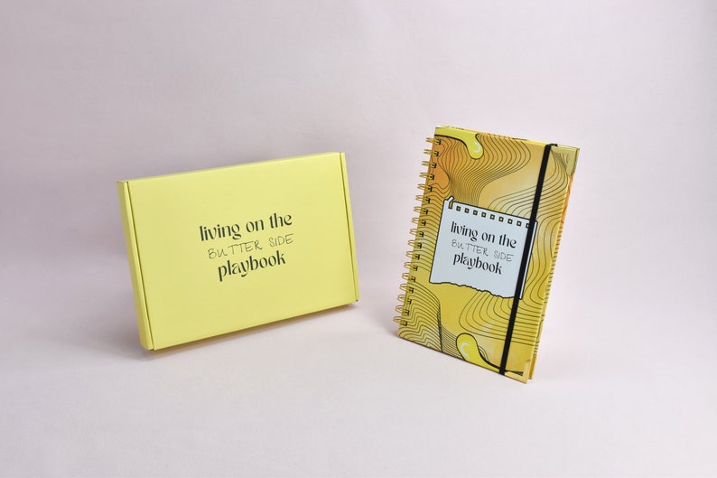 yellow journal, living on the butter side, gold corners and gold binding. black elastic holder. yellow box with brand logo. both items are up right, next to each other.