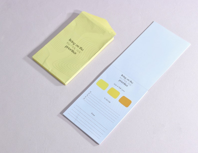 notepad open to display a page. lists acts of self-care, to do list, and notes section. next to the notepad is a  yellow envelope with the brand logo. both items laying down on a surface.