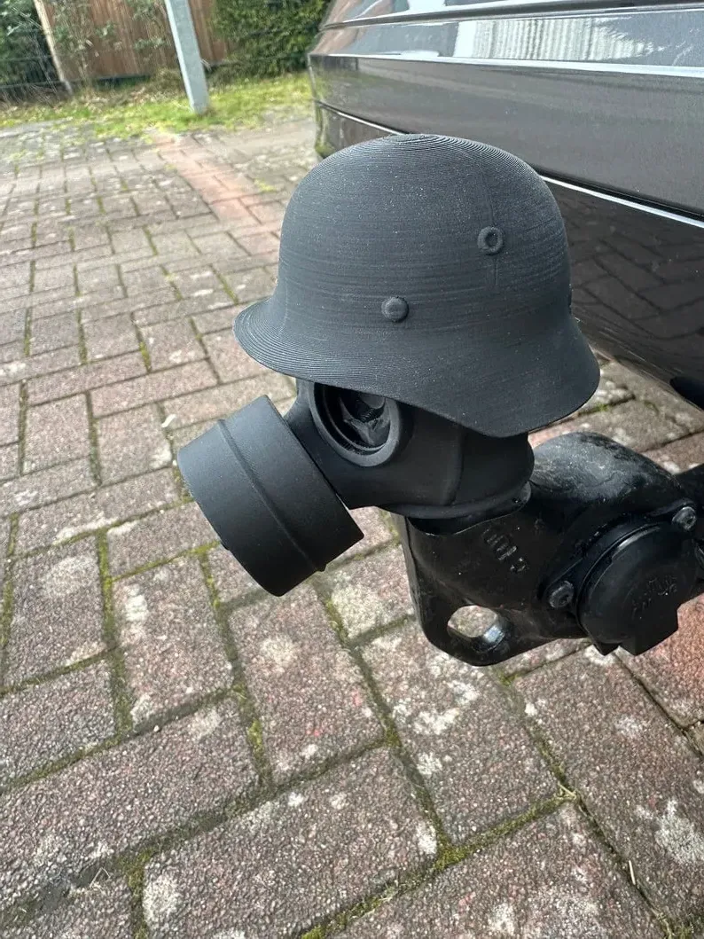 May include: A black gas mask attached to a black helmet. The helmet has a round shape with a brim and is attached to the gas mask with a black fabric covering the connection. The gas mask has a round filter attached to the front.