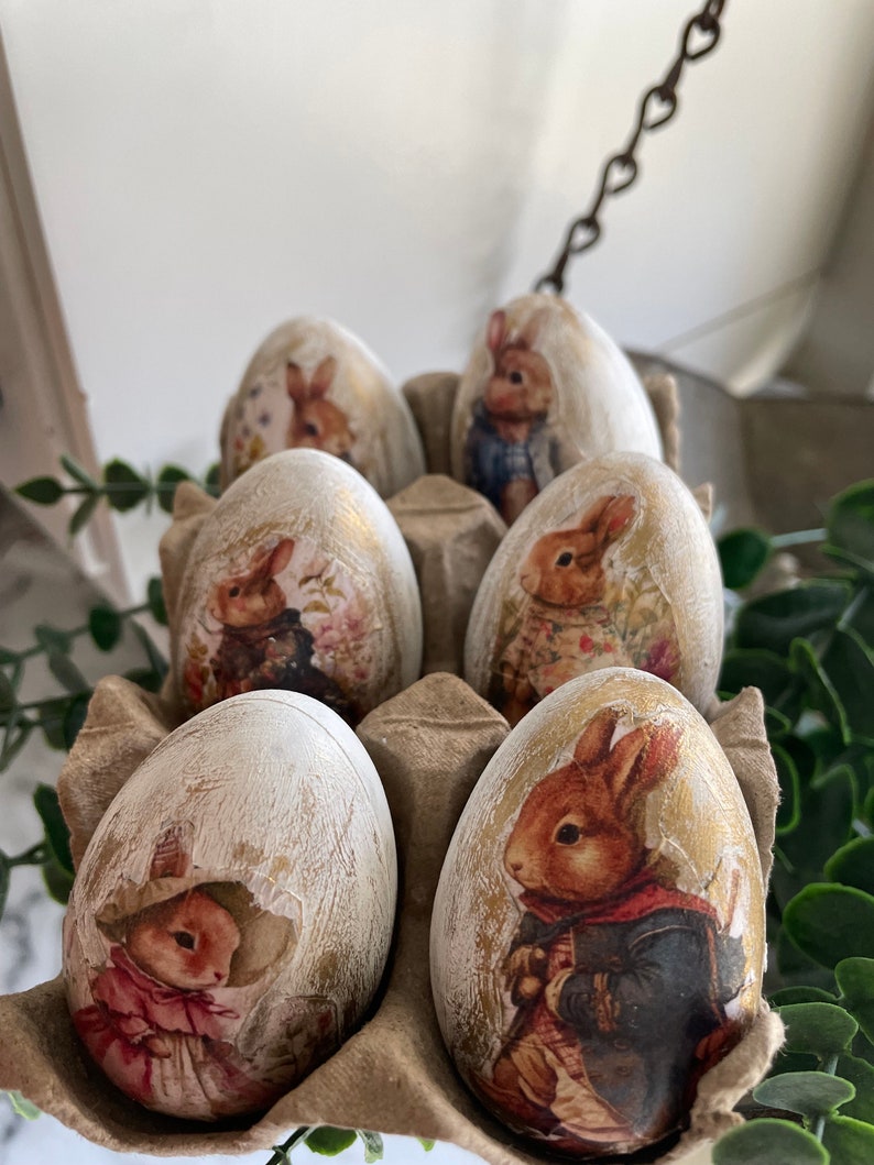 May include: Six decorative Easter eggs in a cardboard egg carton. The eggs are white with a distressed paint finish and feature images of bunnies in various poses. The bunnies are wearing clothing and have a vintage aesthetic.