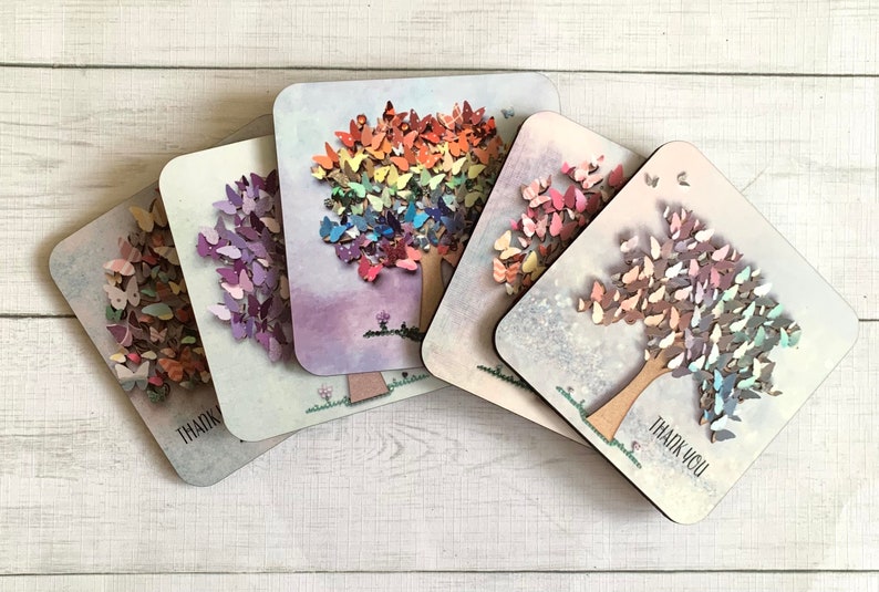 May include: Five square coasters with a tree design made of butterflies. Each coaster has a different color scheme. The coasters have a white background with a light purple, light blue, light pink, and light gray background. The butterflies are made of wood and are painted in various colors. The coasters have the words 'Thank You' printed on them.